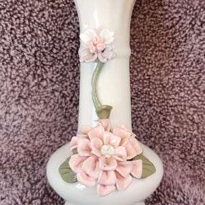 May include: A white ceramic vase with a flared top and a rounded base. The vase is adorned with two clusters of pink and white floral embellishments with green leaves. The vase is approximately 8 inches tall.