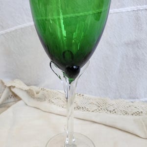 May include: A green-tinted wine glass with a clear stem and base. The bowl of the glass is a deep emerald green, and the stem features a small, dark green sphere. The glass sits on a round, clear base.