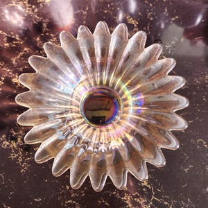 May include: Clear glass bowl with a flower-like design. The bowl has a central, iridescent bubble and petal-shaped edges. The glass reflects light, creating a rainbow effect. The bowl is likely used for decorative purposes or serving.