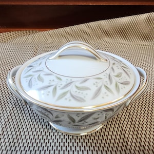 May include: A white porcelain serving dish with a lid, featuring a gold handle and trim. The dish is decorated with a delicate pattern of green and gray leaves. The dish has two side handles.