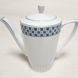 Peut inclure: Cafetière en céramique blanche avec couvercle et anse. Le pot présente un motif géométrique bleu et blanc sur le dessus et une bordure argentée. Le bec verseur est long et fin.