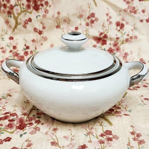 Könnte beinhalten: Eine weiße Zuckerdose aus Keramik mit Deckel und zwei Griffen. Die Schüssel hat einen silbernen Rand um den Rand und den Deckel. Der Hintergrund ist ein Blumenmuster mit rosa und roten Blüten.