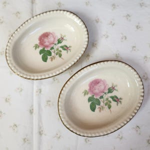 Könnte beinhalten: Zwei ovale, cremefarbene Keramikschalen mit einem Design aus rosa Rosen und grünen Blättern. Die Schalen haben einen goldfarbenen, perlenbesetzten Rand. Der Hintergrund ist ein weißes Blumenmuster.