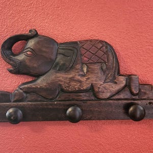 May include: A dark brown wooden wall-mounted hook rack featuring a carved elephant figure. The elephant is depicted in a kneeling position with its trunk raised. The rectangular base includes three rounded wooden pegs for hanging items.