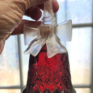 May include: A ruby red bell-shaped glass bottle with a clear, diamond-patterned stopper and a frosted bow detail. The bottle is filled with a deep red liquid. The bell has a diamond pattern.
