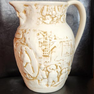 May include: Cream-colored ceramic pitcher with a detailed relief design. The pitcher features a handle and a spout. The relief depicts figures, buildings, and text, including the year "1800". The overall design suggests a historical or vintage theme.