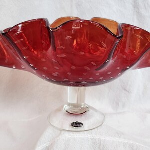 May include: A red glass bowl with a ruffled rim and a clear pedestal base. The bowl has a textured surface with small white dots. The rim has a wavy design, and the glass appears to be translucent.