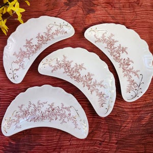 May include: Four white, scalloped-edge serving dishes with a floral design in shades of brown and gold. The dishes are arranged on a red surface, with yellow flowers in the upper left corner.