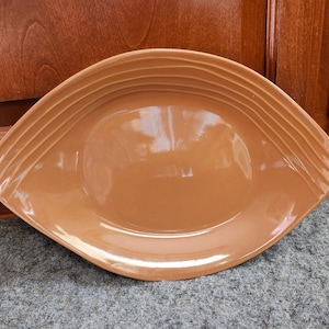 May include: A brown, ceramic serving platter with a unique, eye-shaped design. The platter has a glossy finish and features a series of parallel ridges along the top edge. The overall shape is a rounded rectangle with pointed ends.