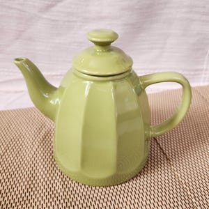 May include: A pale green ceramic teapot with a lid and handle. The teapot has a rounded body with vertical ridges and a curved spout. The teapot is on a brown woven mat.