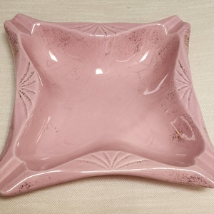 May include: A vintage, square ashtray in a soft pink hue. The ceramic ashtray features a central bowl, four resting areas, and decorative starburst and fan-shaped details. The edges have a distressed, speckled finish.
