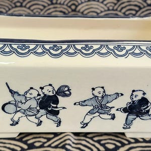 May include: A rectangular ceramic planter with an off-white base and a dark blue rim. The exterior features a repeating blue wave pattern along the top edge and a detailed illustration of five children playing with various items in blue ink.