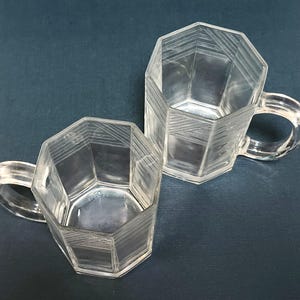 Puede incluir: Dos tazas de vidrio transparente facetadas con asas. Las tazas tienen forma octogonal con diseños lineales grabados. Las asas son redondeadas y transparentes. Las tazas están sobre un fondo azul oscuro.
