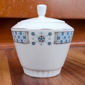 Könnte beinhalten: Weißer Keramik-Zuckertopf mit Deckel, verziert mit einem blau-schwarzen Blumenmuster am Rand. Die Schüssel hat einen goldenen Rand und einen geriffelten Boden. Der Deckel hat einen kleinen, geriffelten Griff.
