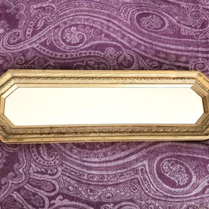 May include: A rectangular, gold-framed mirror with an octagonal shape. The mirror's frame has a decorative, textured design. The mirror is set against a purple paisley patterned background.