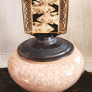 May include: A decorative ceramic jar with a black lid and a wooden topper. The topper features a carved design of fish and Celtic knotwork. The jar has a light pink, crackled glaze finish. The jar is a decorative item.