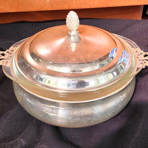 Könnte beinhalten: Eine silberne Servierschale mit Glaseinsatz und Deckel. Der Deckel hat einen dekorativen Knopf und die Schale hat verzierte Griffe. Die Schale ist rund und scheint aus Metall zu sein. Die Schale steht auf einer schwarzen Oberfläche.