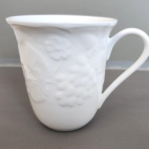 May include: White ceramic mug with a curved handle. The mug features a raised grape and leaf design around the body. The mug has a smooth, glossy finish and a slightly flared rim.