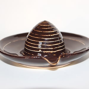 May include: A brown ceramic citrus juicer with a conical reamer and a wide, shallow base. The reamer has horizontal ridges and is surrounded by a circular basin with a spout. The juicer is designed for extracting juice from citrus fruits.