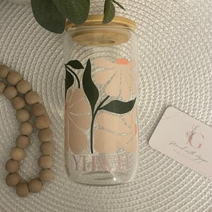Puede incluir: Un vaso de vidrio transparente con tapa de bambú, con un diseño floral en melocotón y verde. La palabra "YHWH" está impresa en el vidrio. También se ve una pequeña tarjeta con gráficos florales.
