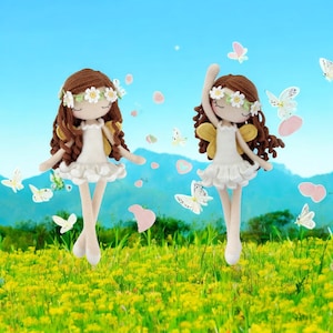 Puede incluir: Dos muñecas de crochet vestidas de hadas con cabello castaño, vestidos blancos y alas amarillas. Están de pie en un campo de flores amarillas con un cielo azul y nubes blancas en el fondo.