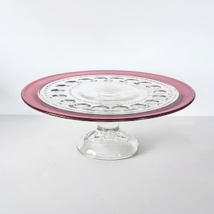May include: A clear glass cake stand with a circular, raised base and a wide, flat top. The rim of the top is a deep pink color. The center of the top has a decorative pattern. The stand is likely used for displaying cakes or other desserts.