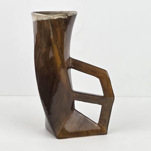 May include: A brown ceramic vase with a unique geometric handle design. The vase has a tall, curved body with a rectangular handle featuring two cut-out shapes. The top rim has a white glaze.