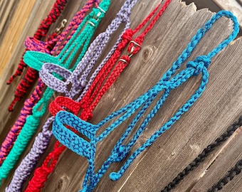 May include: A collection of colorful horse halters in various shades, including red, turquoise, purple, and blue. The halters are made of braided rope and are displayed against a weathered wooden fence. The image showcases the different colors and styles available.