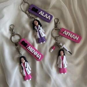 Puede incluir: Llaveros con figuras de médicos en miniatura con batas blancas y trajes rosas o morados. Cada llavero tiene una etiqueta con el texto "DR." seguido de un nombre en letras blancas. Un llavero incluye una jeringa y una pastilla.