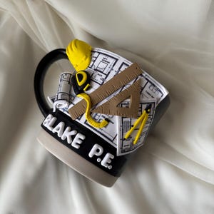 Puede incluir: Una taza de cer&aacute;mica negra y beige con un dise&ntilde;o en 3D de planos arquitect&oacute;nicos, un casco amarillo, una cinta m&eacute;trica, una regla triangular marr&oacute;n y un comp&aacute;s amarillo. El nombre BLAKE P.E. est&aacute; grabado en letras blancas en la base negra.