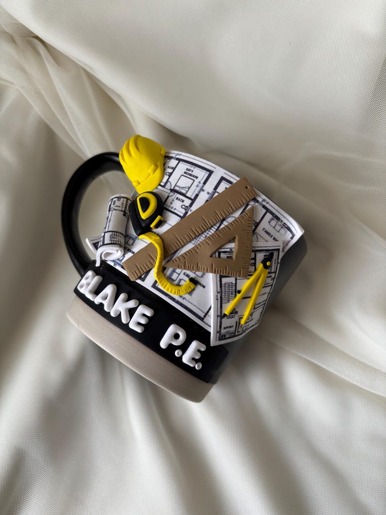 Puede incluir: Una taza de cer&aacute;mica negra y beige con un dise&ntilde;o en 3D de planos arquitect&oacute;nicos, un casco amarillo, una cinta m&eacute;trica, una regla triangular marr&oacute;n y un comp&aacute;s amarillo. El nombre BLAKE P.E. est&aacute; grabado en letras blancas en la base negra.