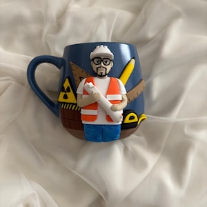 Könnte beinhalten: Eine blaue Keramiktasse mit einem Bauarbeiter-Design. Die Tasse hat einen Henkel und ist mit einem Miniatur-Bauarbeiter mit Helm und Sicherheitsweste verziert, der einen Bauplan hält. Weitere Details sind ein Bleistift, ein Lineal und ein Strahlungssymbol.