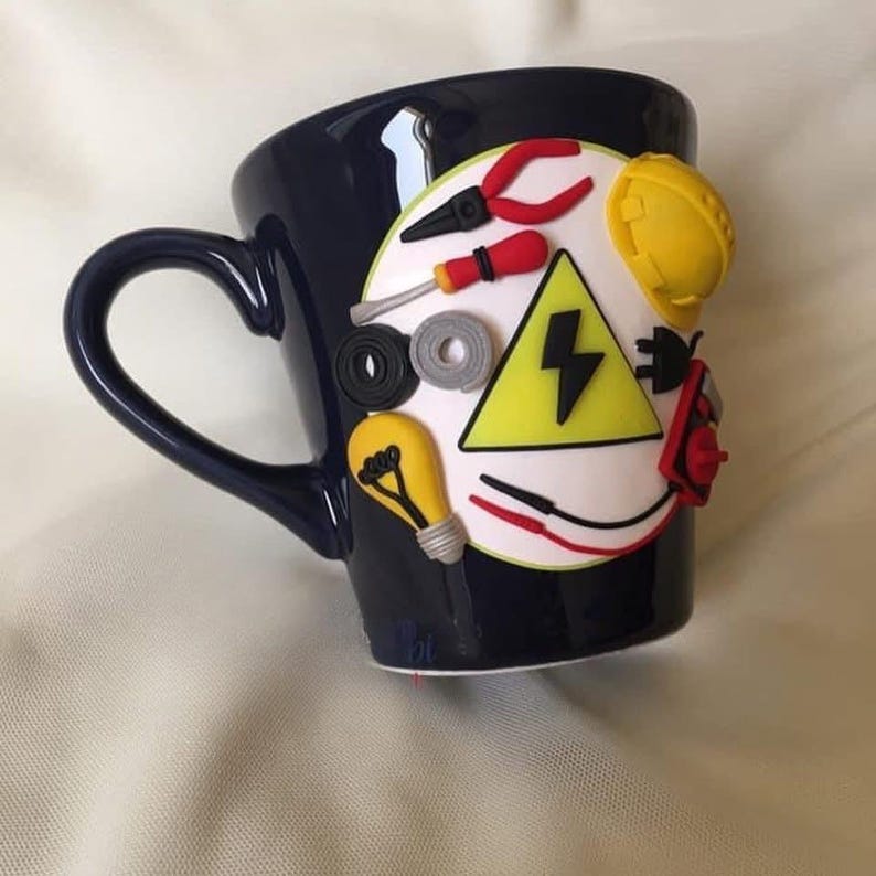 May include: A dark blue ceramic mug featuring a white circular emblem decorated with three-dimensional clay figures of electrical tools, including red pliers, a screwdriver, a yellow hard hat, a light bulb, and a yellow triangle with a black bolt.