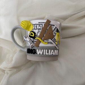 Puede incluir: Una taza de cer&aacute;mica gris claro con un dise&ntilde;o decorativo en 3D de herramientas de ingenier&iacute;a, incluyendo un casco amarillo, una cinta m&eacute;trica, una regla, una escuadra y un comp&aacute;s sobre un plano. El nombre ENG. WILIAM est&aacute; en relieve blanco.