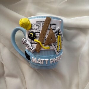 Puede incluir: Una taza azul claro decorada con herramientas de arquitectura en arcilla 3D, incluyendo un casco amarillo, una cinta m&eacute;trica, una regla, un cartab&oacute;n, un comp&aacute;s y planos enrollados. La taza presenta el texto en relieve blanco MATT PHELPS.
