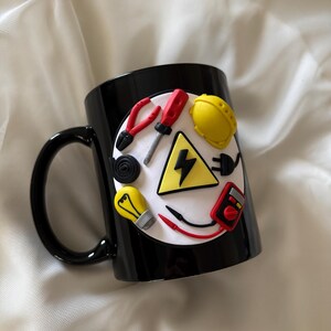 May include: A black ceramic mug featuring a circular white emblem decorated with three-dimensional clay figures of an electrician's tools, including a yellow hard hat, a red screwdriver, pliers, a light bulb, a multimeter, and a lightning bolt.