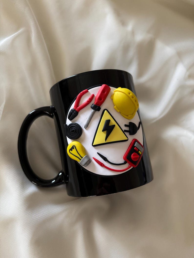 May include: A black ceramic mug featuring a circular white emblem decorated with three-dimensional clay figures of an electrician's tools, including a yellow hard hat, a red screwdriver, pliers, a light bulb, a multimeter, and a lightning bolt.