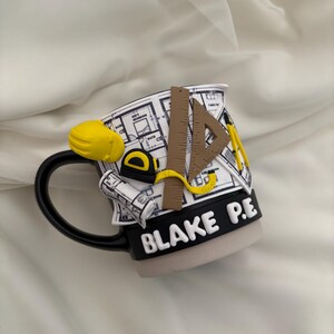 Puede incluir: Una taza de cer&aacute;mica con asa y base negras, con una superficie blanca decorada con un patr&oacute;n de planos. Los elementos 3D incluyen un casco amarillo, una cinta m&eacute;trica, una regla marr&oacute;n, una escuadra y el texto en blanco BLAKE P.E.