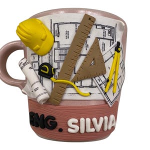 Puede incluir: Una taza de cer&aacute;mica rosa claro con un dise&ntilde;o arquitect&oacute;nico en 3D que incluye un casco amarillo, un plano enrollado, una cinta m&eacute;trica, una regla marr&oacute;n, una escuadra y un comp&aacute;s amarillo. El texto ENG. SILVIA est&aacute; en relieve en letras.