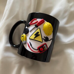 May include: A black ceramic mug featuring a circular white emblem decorated with three-dimensional miniature electrical tools and symbols, including a yellow hard hat, a light bulb, a multimeter, pliers, a screwdriver, and a black lightning bolt.