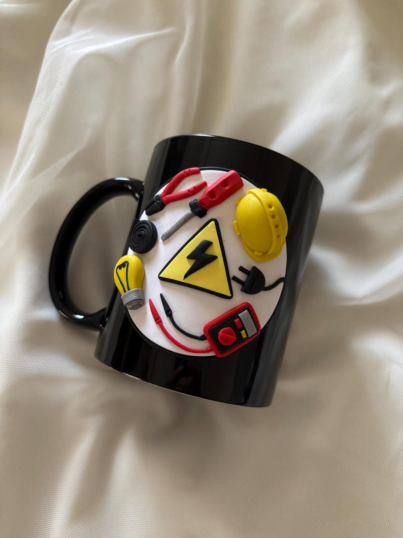May include: A black ceramic mug featuring a circular white emblem decorated with three-dimensional miniature electrical tools and symbols, including a yellow hard hat, a light bulb, a multimeter, pliers, a screwdriver, and a black lightning bolt.