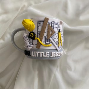 Puede incluir: Una taza de cer&aacute;mica blanca con un dise&ntilde;o arquitect&oacute;nico en 3D que incluye un casco amarillo, una cinta m&eacute;trica, una regla, una escuadra y un comp&aacute;s. La taza muestra planos y el texto LITTLE JESSY en letras blancas sobre una base gris.