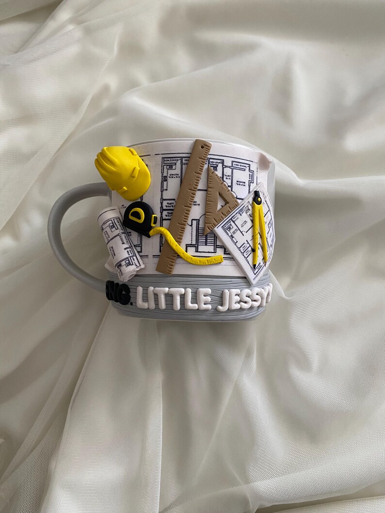 Puede incluir: Una taza de cer&aacute;mica blanca con un dise&ntilde;o arquitect&oacute;nico en 3D que incluye un casco amarillo, una cinta m&eacute;trica, una regla, una escuadra y un comp&aacute;s. La taza muestra planos y el texto LITTLE JESSY en letras blancas sobre una base gris.