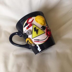 May include: A dark blue ceramic mug featuring a white circular applique decorated with three-dimensional polymer clay shapes including red pliers, a yellow hard hat, a yellow triangle with a black lightning bolt, a lightbulb, and electrical tools.
