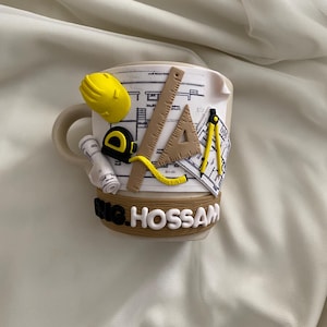 May include: A beige ceramic mug decorated with 3D clay architectural elements including a yellow hard hat, a tape measure, a ruler, a triangle, a compass, and rolled blueprints. The base features a brown wood-textured band with the text ENG. HOSSAM.