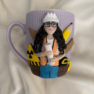 May include: A lilac ceramic mug decorated with a sculpted female construction worker. She wears a white hard hat, glasses, and an orange safety vest. The mug is adorned with a pencil, ruler, and other construction-related elements.