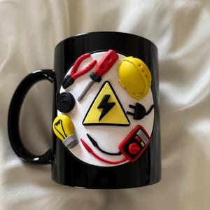 May include: A black ceramic mug featuring a circular white emblem decorated with three-dimensional clay figures of electrical tools, including a yellow hard hat, a red screwdriver, pliers, a light bulb, a multimeter, and a yellow high-voltage sign.