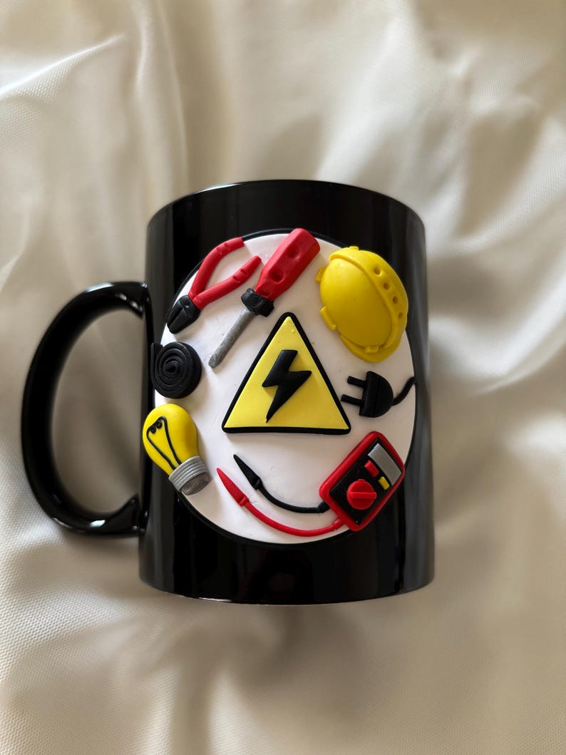 May include: A black ceramic mug featuring a circular white emblem decorated with three-dimensional clay figures of electrical tools, including a yellow hard hat, a red screwdriver, pliers, a light bulb, a multimeter, and a yellow high-voltage sign.