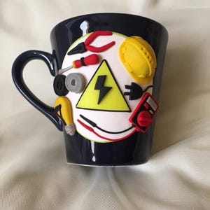 May include: A dark blue ceramic mug featuring a white circular emblem decorated with three-dimensional clay figures of a yellow hard hat, a red multimeter, a yellow light bulb, red pliers, a screwdriver, electrical tape, and a yellow lightning bolt.