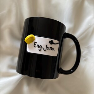 May include: A glossy black ceramic mug featuring a white rectangular plaque on the front. The plaque displays the text Eng. Jana in black script, accompanied by a small yellow hard hat icon and a black electrical plug icon attached to the text.